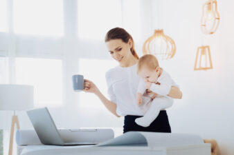 woman holding a baby and coffee mug while looking at laptop