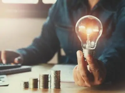 man holding up a lightbulb while looking at stack of coins