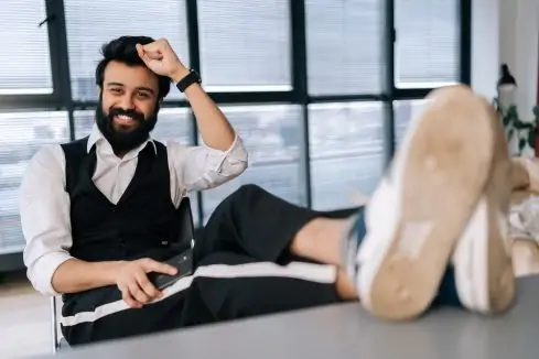 Portrait of cheerful bearded businessman sitting at office desk with feet up on table with laptop, finished work, job done, completed all tasks, relaxing after hard workday, smiling looking at camera.